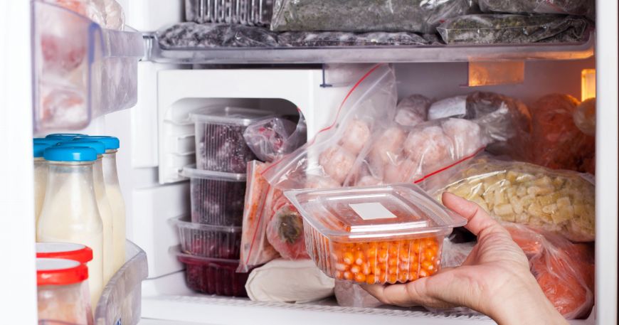 A person removes a tub of food from a freezer. The tub, along with other items in the freezer,  has been labelled with white freezer labels.