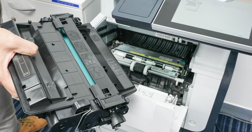 A person holding a toner cartridge in front of an office laser printer; laser printers are a type of hot fuse printer system.