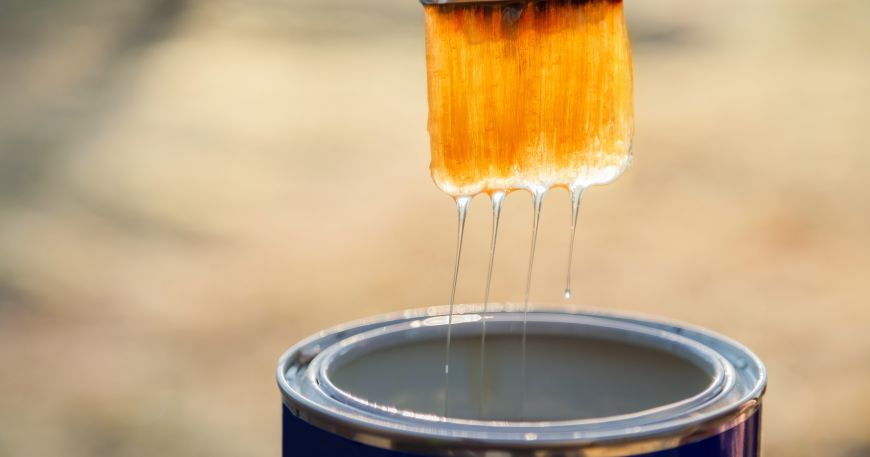A paintbrush that has been dipped in a can of lacquer; the brush is being held above the can as the lacquer drips off the end of the brush.