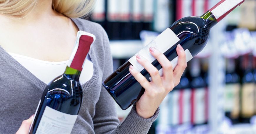 A person looking at two bottles of wine; each bottle has a primary label (on the front of the bottle) and a secondary label (on the back).