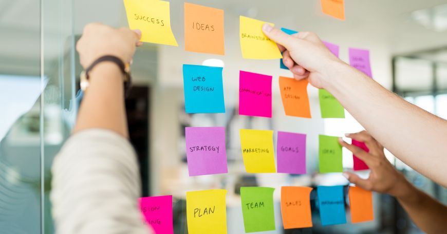 People moving around a set of post it notes on a glass wall; a repositionable label can be stuck onto one item or surface and then moved to another item or a different position.