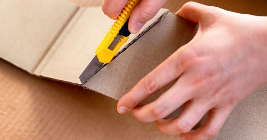 A person uses a retractable knife to score a piece of cardboard. Scoring means cutting partway through a material to make it easier to manipulate; here the person has cut through the top layers of the cardboard, leaving the bottom layer intact.