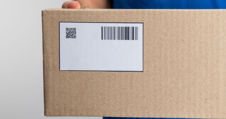 A person holding a labelled cardboard box; the label is a square cut corner rectangle, which means that its edges meet in a pointed corner at a 90° angle.