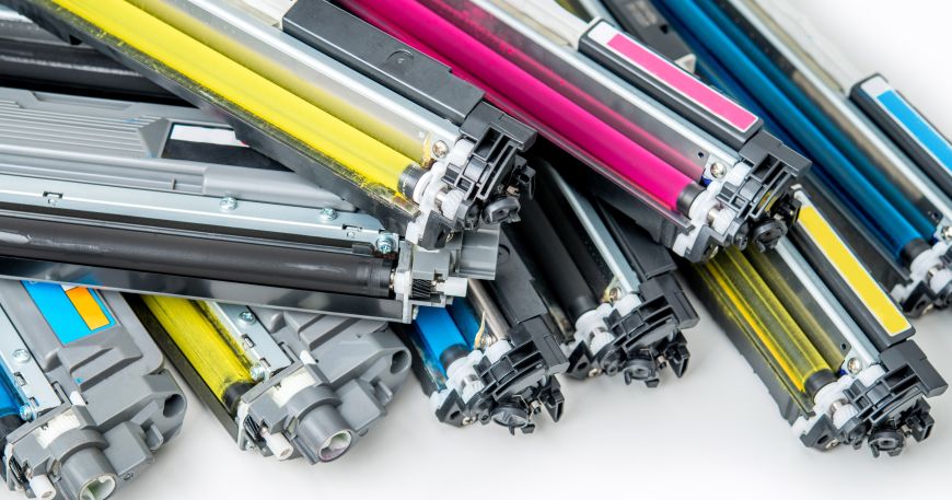 A selection of laser toner cartridges in the four CMYK colours (cyan, magenta, yellow, and black); loose toner, a powder, can be seen on the roller of each cartridge. 