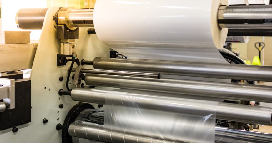 A roll of top lamination on a printing press; this clear film will be applied onto materials as a protective and/or decorative layer during the manufacturing process.