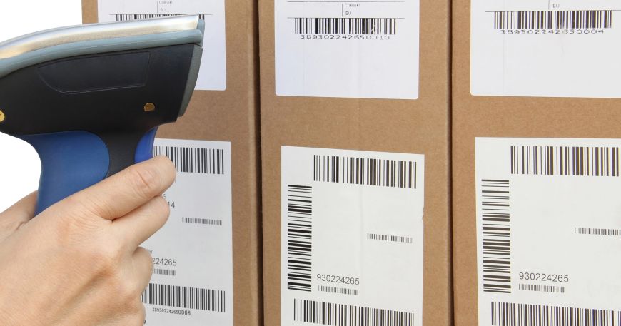 A person holds a barcode scanner next to three boxes that are all labelled with tracking labels; a tracking label is used to track an item throughout the supply chain including manufacturing, distribution, storage, and delivery.
