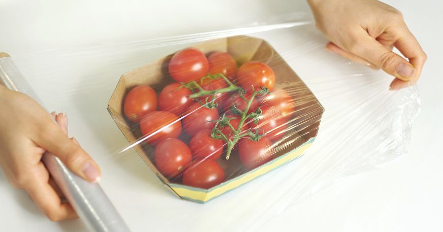 A person covers a box of tomatoes with a transparent film; the transparent film can be seen through, which means the food remains visible below this protective layer.