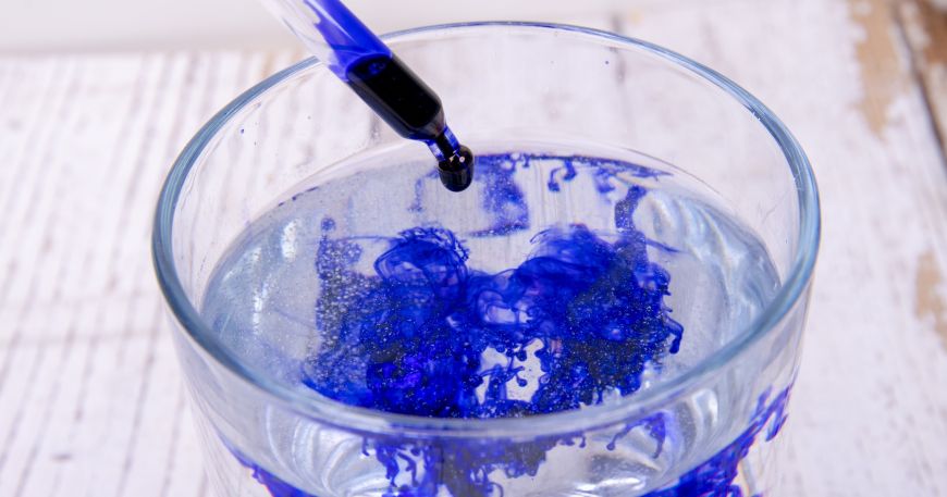 A pipette is used to add drops of a blue liquid into a glass containing a clear vehicle; the vehicle is the liquid component in inks, which holds the colourant and binds it to the substrate.
