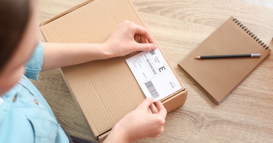 A woman applies an address label onto a parcel. Application describes both the purpose of the label (to add address, delivery, and tracking information to the parcel) and the process of sticking the label onto the box.