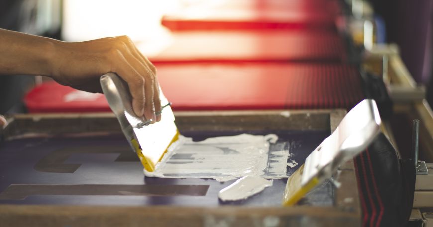 A person uses screen printing to add a design to a t-shirt. A template has been lain over the screen and a squeegee is used to force the ink through the screen in the correct places to form the required design.