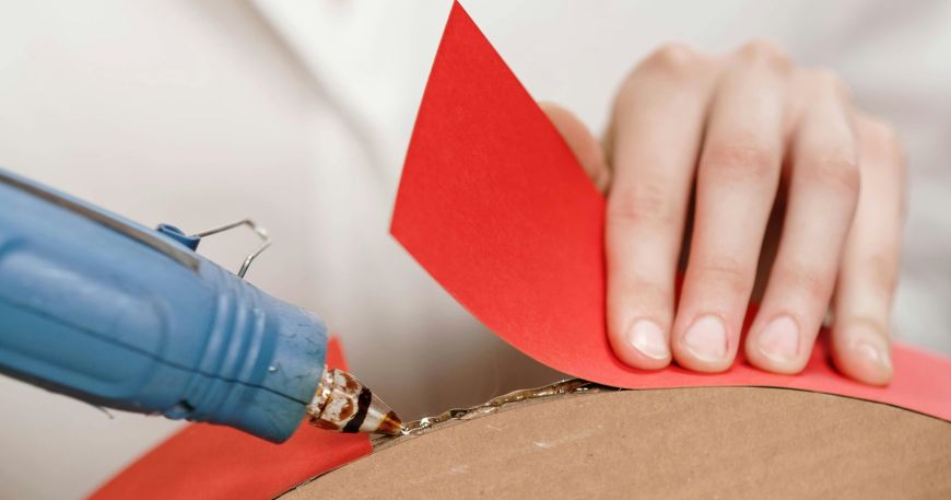 A person bonds a strip of red card onto cardboard using a glue gun.