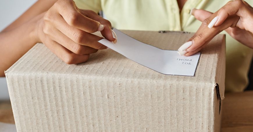 A woman applies a label onto a cardboard box; both the white paper face material of the label and the cardboard box can be described as an adherend.