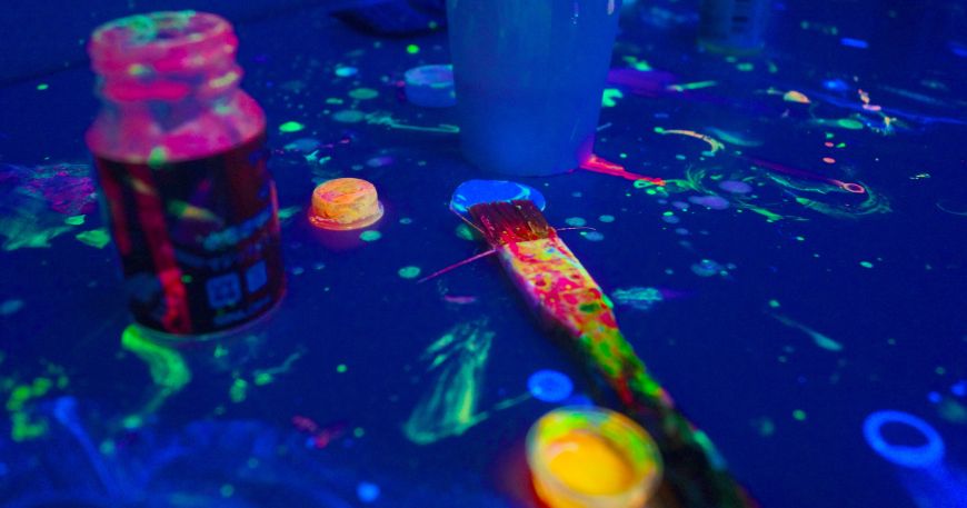 A table spattered with fluorescent paint; there are pots and buckets containing fluorescent paint on the table, along with a paintbrush. 