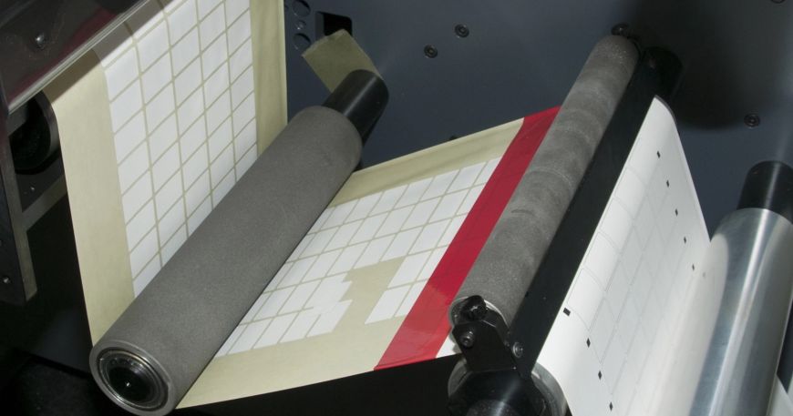 A set of labels being processed through a printing machine; a number of labels have delaminated from the backing sheet.
