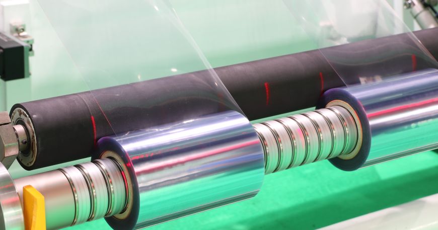 Transparent rolls of film loaded onto a machine for slitting large rolls into narrower rolls.