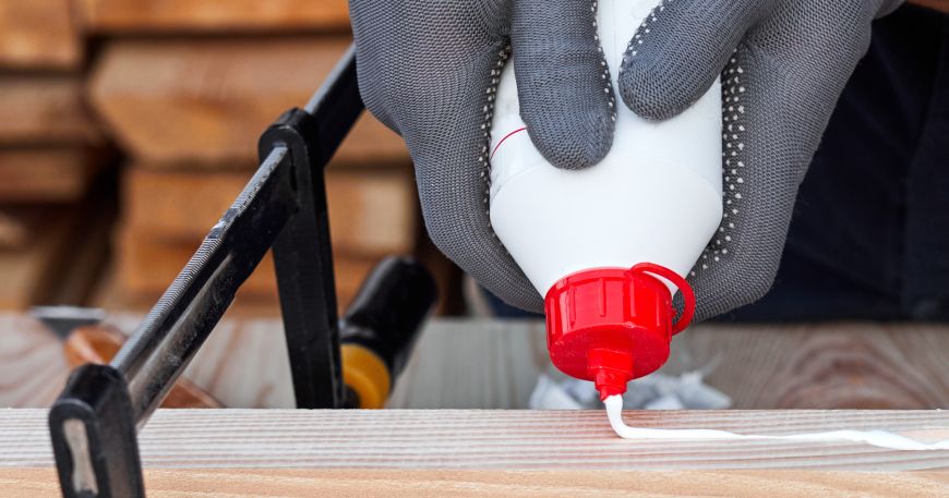 A person applies wood glue onto wooden sections of a table; many wood glues are water based adhesives, which allows them to be spread in liquid form across wooden surfaces.