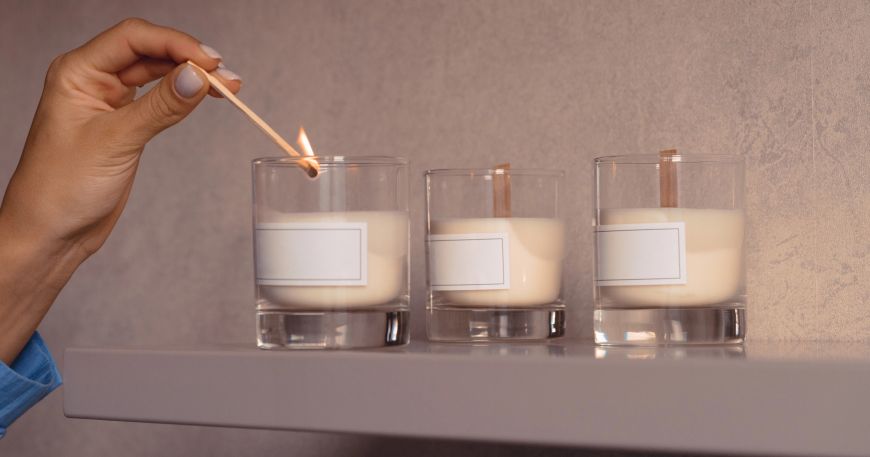 A person lights the first of three candles on a shelf; each candle is labelled with a white label. Acrylic based adhesives are popular for product labels like those used to label the glass candle holders in this image.