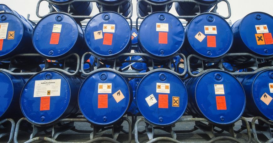 Several blue chemical barrels stacked on metal racking; each barrel is labelled with a number of labels. Chemical barrels are often labelled using marine adhesives to ensure that labels remain in place even if a barrel ends up in sea water.