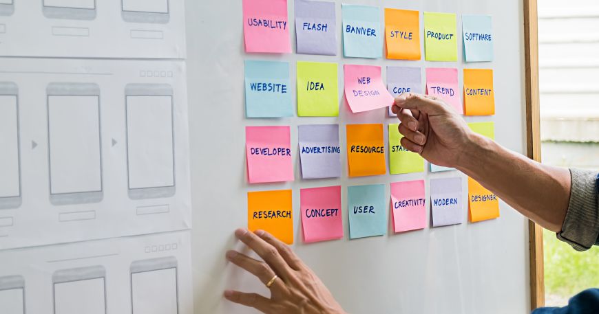 A person removes a post it note from a white board covered in notes. Post it notes may be made with microsphere adhesives because this allows them to be removed and repositioned without damaging surfaces.