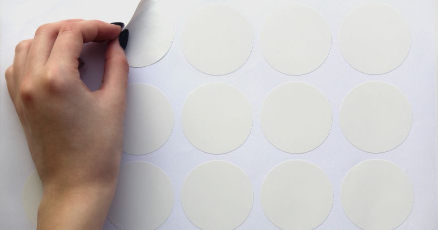 A person peels a round label off a sheet of pressure sensitive labels. 