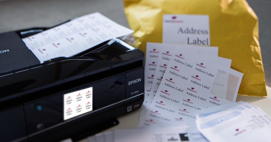 A desk with a printer, sheets of printed address labels, and envelopes and packages labelled with address labels.