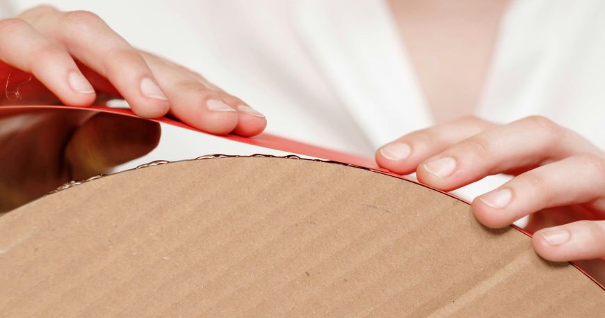 A woman uses glue to adhere a strip of decorative red material onto a cardboard base. 