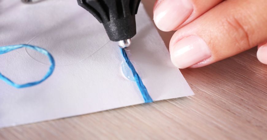 A glue gun is used to stick blue ribbon onto white card. The ribbon can be moved for a short time until the glue dries into its ultimate adhesion.