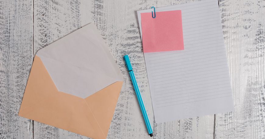 A brown C5 envelope lying next to an A4 sheet of paper with a sheet of pink note paper paper clipped to the top with a blue pen in between the two items.