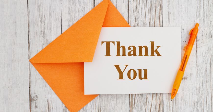 An orange C6 envelope lying on a wooden desk; a small Thank You card and orange pen lie next to the envelope.